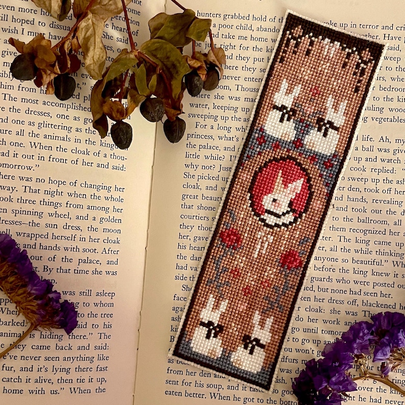 May include: A brown bookmark with a cross-stitch design featuring three white rabbits with black eyes and noses. The rabbits are surrounded by red, blue, and green flowers. The bookmark has the text "The Little Rabbit" in white lettering.