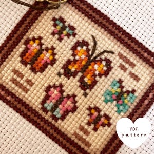 Peut inclure: Motif de point de croix avec un cadre brun sur fond blanc et cinq papillons colorés. Les papillons sont orange, rose et bleu. Le texte "PDF pattern" est dans un cœur blanc en bas à droite.