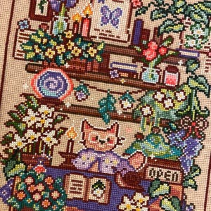 May include: A cross-stitch piece depicting a cozy scene with a cat reading a book. The artwork features shelves with potted plants, candles, and a sign that says "OPEN". The color palette includes shades of brown, purple, green, and yellow.