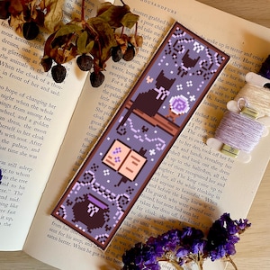 Könnte beinhalten: Lilafarbener und schwarzer Pixel-Art-Lesezeichen mit einer schwarzen Katze, einem Kessel, einem Buch und einer Blume. Das Lesezeichen hat einen sternenklaren Nachthimmel als Hintergrund mit einem weißen Rand.