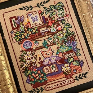 May include: Cross-stitch art piece featuring a cat in a purple vest, surrounded by plants and books. The artwork includes the text "The Herbalist" and "OPEN". The color palette includes shades of beige, green, purple, and orange, with a gold frame.