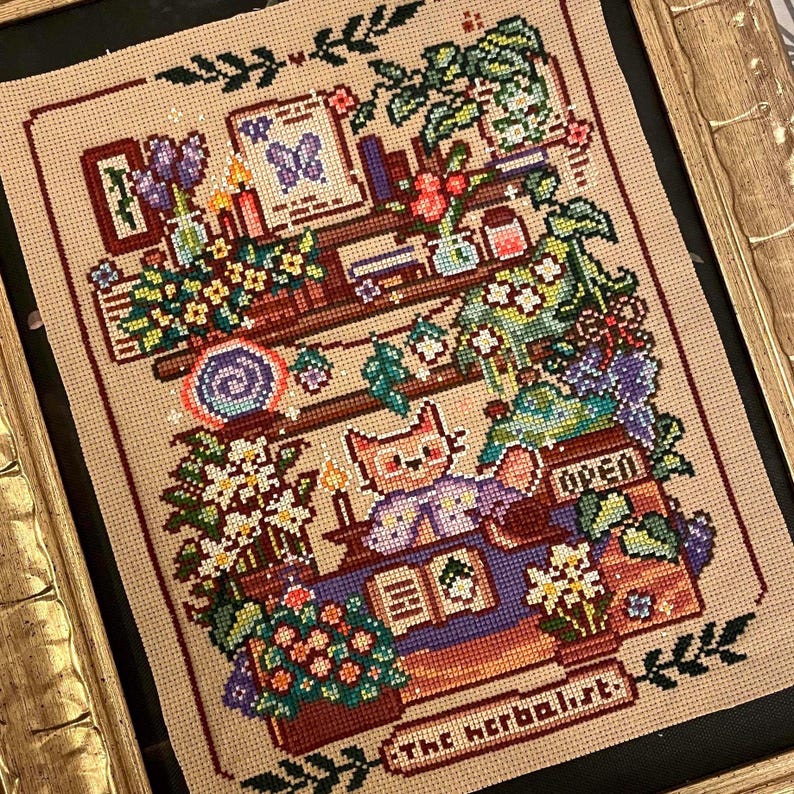May include: Cross-stitch art piece featuring a cat in a purple vest, surrounded by plants and books. The artwork includes the text "The Herbalist" and "OPEN". The color palette includes shades of beige, green, purple, and orange, with a gold frame.