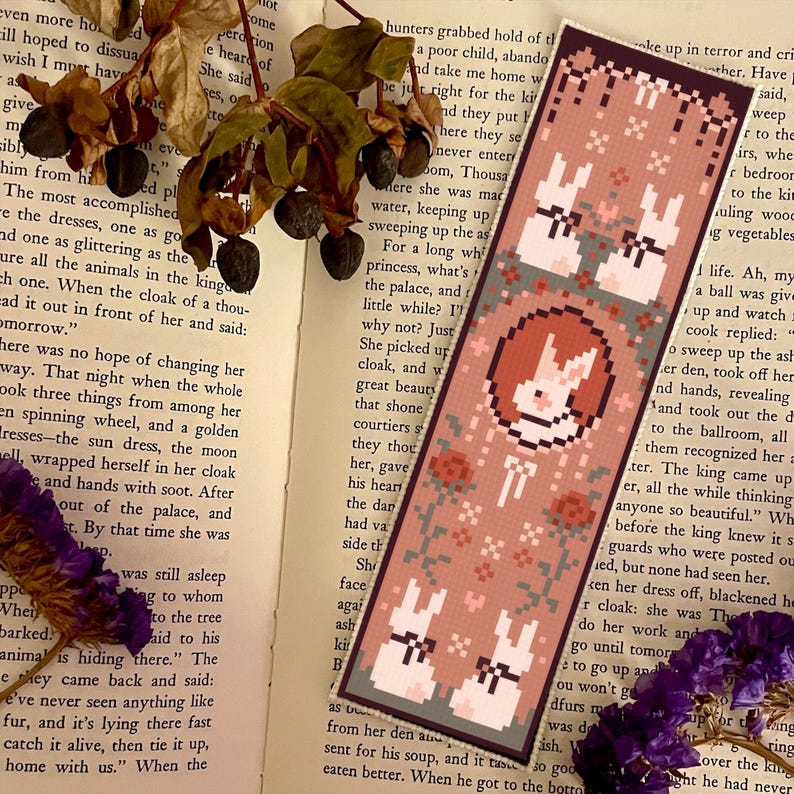 May include: A pink and green bookmark with a pixelated design of three white rabbits with bows. The rabbits are on a pink background with green vines and flowers. The bookmark has the text "Just a little while?" on the left side.