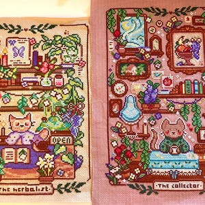 May include: Two cross-stitch artworks. The left piece, "The Herbalist," features a cat behind a counter with plants and books. The right piece, "The Collector," shows a cat with a collection of objects. Both are framed.
