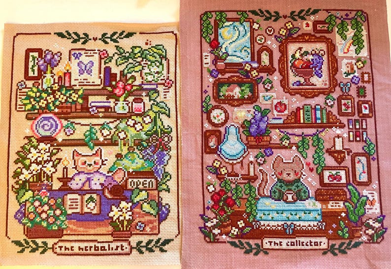 May include: Two cross-stitch artworks. The left piece, "The Herbalist," features a cat behind a counter with plants and books. The right piece, "The Collector," shows a cat with a collection of objects. Both are framed.