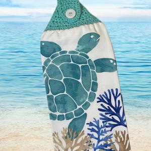 Puede incluir: Una toalla de cocina de color turquesa y blanco con un borde de ganchillo y un botón. La toalla presenta un diseño de tortuga marina y coral.