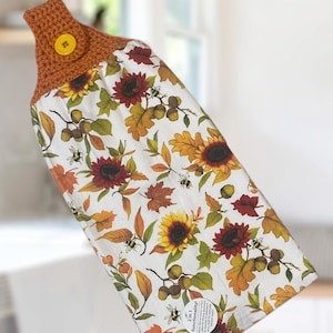 May include: A hanging kitchen towel with a crochet top and a button closure. The towel features a white background with a fall-themed pattern of sunflowers, leaves, acorns, and bees. The crochet top is a warm orange color.