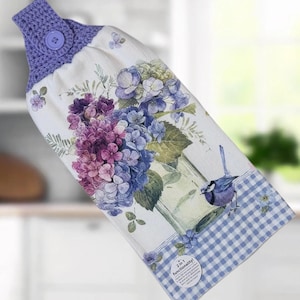 May include: A hanging kitchen towel featuring a purple crocheted top with a matching button. The white fabric displays a floral print of blue and purple hydrangeas in a glass vase, a small blue bird, and a blue gingham border with a circular label.