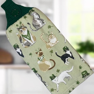 May include: A green and white kitchen towel with a pattern of cats wearing St. Patrick's Day hats and shamrocks. The top of the towel is a dark green crochet loop.