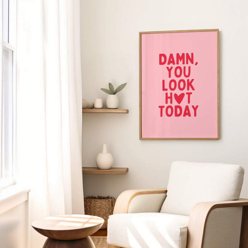 May include: A pink framed print with the text "DAMN, YOU LOOK HOT TODAY" in red with a red heart in the word "HOT". The print is hanging on a white wall in a room with a white armchair and a wooden side table.