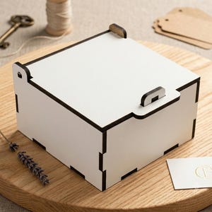 Könnte beinhalten: Eine weiße Holzbox mit schwarzen Rändern und einem Klappdeckel. Die Box steht auf einer Holzoberfläche mit Lavendel, einem Schlüssel und einer Garnrolle. Eine kleine Karte mit einem goldenen Buchstaben ist ebenfalls vorhanden.