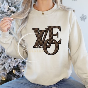 Op de afbeelding: Een beige sweatshirt met een bruin en wit geruit patroonontwerp dat "WE" in grote letters spelt.