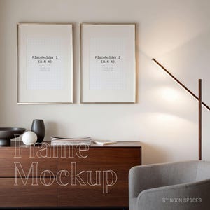 May include: Two gold picture frames with white mats and a white background hanging on a light beige wall. The frames are next to each other. The text "Placeholder 1 (DIN A)" is in the top left frame and the text "Placeholder 2 (DIN A)" is in the top right frame. A brown wooden dresser with two drawers is in the bottom center of the image. A gray armchair is in the bottom right corner of the image. A floor lamp with a long, thin, horizontal arm is in the right side of the image. The text "Frame Mockup" is in white letters on the dresser. The text "BY NOON SPACES" is in white letters on the bottom right corner of the image.