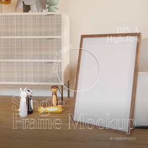 May include: A brown wooden frame mockup with a white background. The frame is leaning against a white wall and a light brown wooden cabinet. The frame is surrounded by a light brown wooden floor. The text "DIN A High-Res Single Frame Mockup" is written on the floor in front of the frame. The text "BY NOON SPACES" is written below the frame.