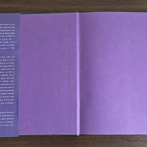 K&ouml;nnte beinhalten: Aufgeschlagenes Kochbuch mit lila Innenseiten und einem dunkelvioletten Buchr&uuml;cken. Die linke Seite hat wei&szlig;en Text, die rechte Seite ist leer. Das Buch liegt auf einer Holzoberfl&auml;che.