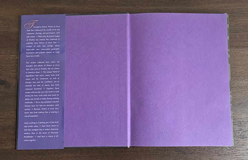 K&ouml;nnte beinhalten: Aufgeschlagenes Kochbuch mit lila Innenseiten und einem dunkelvioletten Buchr&uuml;cken. Die linke Seite hat wei&szlig;en Text, die rechte Seite ist leer. Das Buch liegt auf einer Holzoberfl&auml;che.