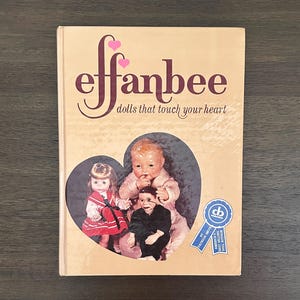 May include: A hardcover book titled Effanbee: dolls that touch your heart with a tan cover. The front features a heart-shaped cutout displaying three dolls and a blue circular seal with the text Patricia Smith America's Best Selling Doll Author.