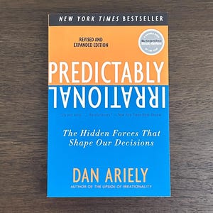 May include: A hardcover book titled "Predictably Irrational" by Dan Ariely. The book cover features orange and blue sections with white text. The book is a New York Times Bestseller, revised and expanded edition, with a "Notable Book" seal.