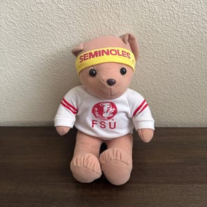 May include: A plush teddy bear wearing a yellow headband with "SEMINOLES" printed on it. The bear is dressed in a white shirt with red stripes on the sleeves and the letters "FSU" on the front. The bear is sitting on a wooden surface.