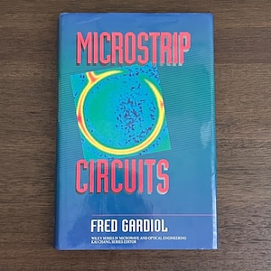 May include: A book titled "MICROSTRIP CIRCUITS" by Fred Gardiol. The cover is blue with the title in red and a graphic of a yellow and green circle. The book is part of the Wiley Series in Microwave and Optical Engineering.