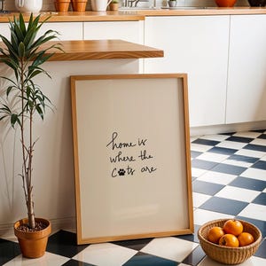 May include: A framed print with the text "home is where the cats are" in a kitchen setting. The print is leaning against a wall next to a potted plant and a basket of oranges. The floor has a black and white checkered pattern.
