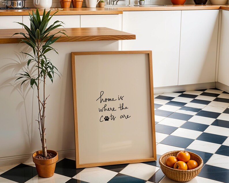 May include: A framed print with the text "home is where the cats are" in a kitchen setting. The print is leaning against a wall next to a potted plant and a basket of oranges. The floor has a black and white checkered pattern.
