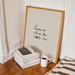 May include: A framed print with the text "home is where the cats are" in black script. The print leans against a wall next to a stack of books, a white mug with coffee, and a striped rug. The frame is made of light wood.