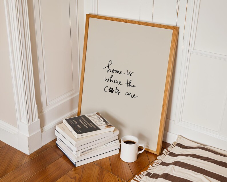 May include: A framed print with the text "home is where the cats are" in black script. The print leans against a wall next to a stack of books, a white mug with coffee, and a striped rug. The frame is made of light wood.