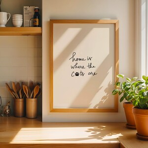 May include: A framed print with the text "home is where the cats are" in a kitchen setting. The print is in a light wood frame. Two potted plants sit on a wooden countertop near a window.