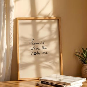 May include: A framed print with the text "home is where the cats are" in a handwritten style. The print is in a light wood frame and is displayed on a wooden surface. A small potted plant and books are also visible.