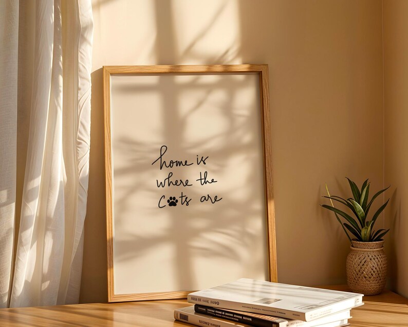 May include: A framed print with the text "home is where the cats are" in a handwritten style. The print is in a light wood frame and is displayed on a wooden surface. A small potted plant and books are also visible.