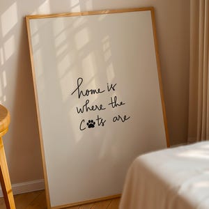 May include: A framed print with the text "home is where the cats are" in black script. The print has a white background and a wooden frame. The image is leaning against a wall next to a wooden stool and a bed.