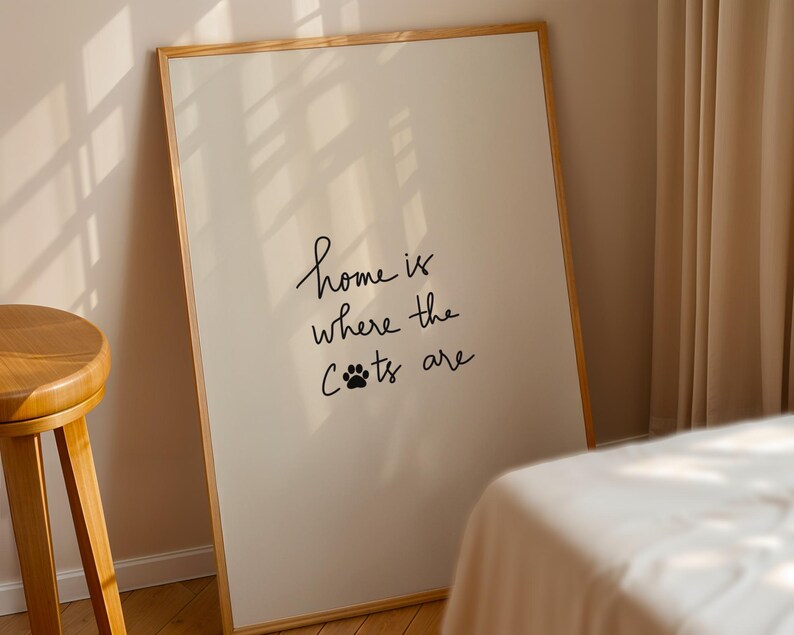 May include: A framed print with the text "home is where the cats are" in black script. The print has a white background and a wooden frame. The image is leaning against a wall next to a wooden stool and a bed.