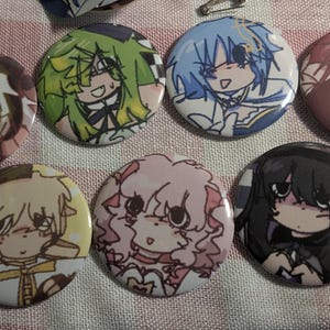Puede incluir: Una colección de siete botones redondos con personajes de estilo anime. Cada botón presenta un diseño de personaje diferente con varios colores de cabello, incluyendo verde, azul, rosa y negro. Los botones están dispuestos sobre una superficie de tela estampada.