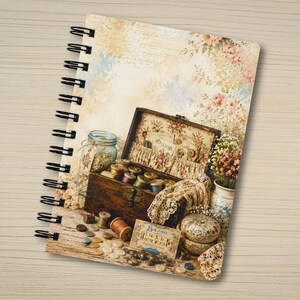 May include: A spiral-bound notebook featuring a printed illustration of a wooden sewing box filled with spools of thread, lace, buttons, and a pincushion. The background shows a soft floral pattern with script text and a glass jar of buttons.