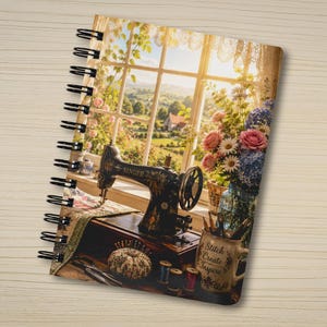 May include: A spiral-bound notebook featuring a printed image of a vintage black Singer sewing machine on a wooden table. Beside it sits a pincushion, thread spools, and a mug with the text Stitch Create Inspire, all set before a sunny window.