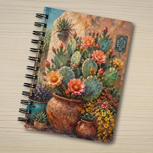 May include: A spiral-bound notebook featuring a vibrant painting of green prickly pear cacti with orange and pink blossoms in terracotta pots. The artwork depicts a sunlit wall, small potted plants, and yellow flowers, all rendered in a textured, painterly style.