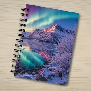 May include: A spiral-bound notebook featuring a vibrant cover illustration of a snow-covered mountain range reflected in a calm lake under a bright aurora borealis sky with shades of purple, blue, and green, and frost-covered trees in the foreground.