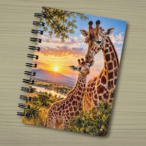 May include: A spiral-bound notebook featuring a vibrant cover illustration of an adult giraffe and a calf standing together in a savanna landscape at sunset, with a river, distant mountains, and golden light filling the background scene.