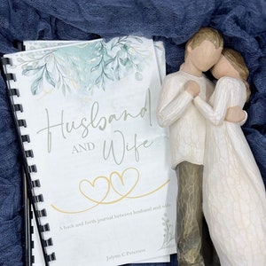 May include: A white journal with a blue and green floral design on the cover. The cover reads "Husband and Wife" and "A back and forth journal between husband and wife". The journal is on a blue fabric surface with two gold rings and a pair of white figurines of a couple embracing.
