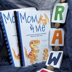 May include: Two blue and white journals with the text "Mom & Me" and "A Keepsake Journal Between Mother and Son". The journals are next to a set of colorful wooden blocks that spell out "RAW" with a red, white, and blue block with the letter "R" in the foreground.