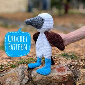 Puede incluir: Patrón de crochet para un juguete amigurumi de alcatraz de patas azules. El juguete es blanco con alas marrones y patas azules. El juguete está sentado en una roca.