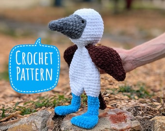 May include: Crochet pattern for a blue-footed booby amigurumi toy. The toy is white with brown wings and blue feet. The toy is sitting on a rock.