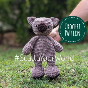 Könnte beinhalten: Ein gehäkelter Koala-Bär in Grau- und Brauntönen, mit kleinen Ohren und einer runden Nase. Das Stofftier wird vor einem grünen Hintergrund gehalten. Der Text auf dem Bild lautet "Crochet Pattern" und "#ScattaYourWorld".