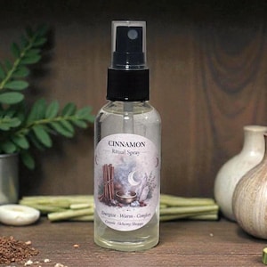 May include: A clear glass spray bottle with a black cap and a label that reads "CINNAMON Ritual Spray." The label features a bundle of cinnamon sticks, a bowl, and a crescent moon design. The bottle is filled with a clear liquid.