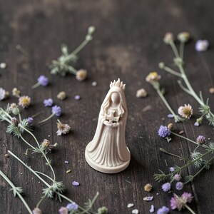 Puede incluir: Figura beige de una figura vestida con una corona, sosteniendo un objeto. La figura est&aacute; rodeada de peque&ntilde;as flores y vegetaci&oacute;n sobre una superficie de madera oscura. La figura mide unos 7 cm de altura.