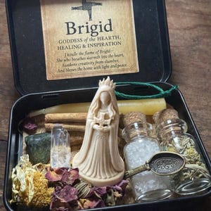 Puede incluir: Una caja met&aacute;lica negra que contiene una estatua de la diosa Brigid, velas, cristales, flores secas y peque&ntilde;os frascos de vidrio. La caja tiene una etiqueta con texto sobre Brigid, la diosa del hogar, la curaci&oacute;n y la inspiraci&oacute;n.