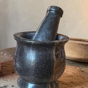 May include: A dark gray mortar and pestle set. The mortar is a rounded bowl with a flared rim and a pedestal base. The pestle is cylindrical with a rounded handle. The set is made of stone and sits on a wooden surface.