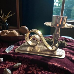 May include: A brass candlestick holder with a decorative scroll design and a round candle cup. The base is rectangular with a diamond pattern. The candlestick is on a burgundy tablecloth.