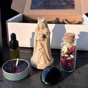 May include: An assortment of spiritual items, including a beige goddess statue holding an apple, a small glass bottle with a cork, a black obsidian stone, a small candle, and a dropper bottle. The items are arranged on a wooden surface.
